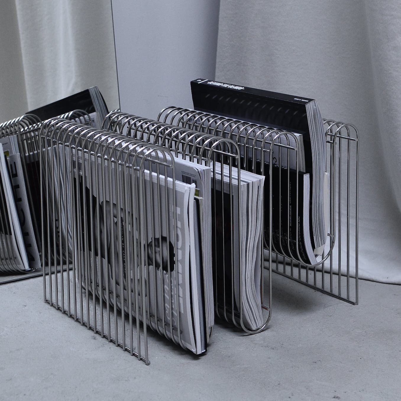 Collection of modern fashion magazines on metal rack placed on floor near mirror and curtain in room corner