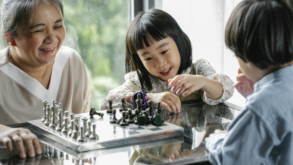 Crop cheerful elderly ethnic female helping adorable little grandchildren playing chess at table in light room