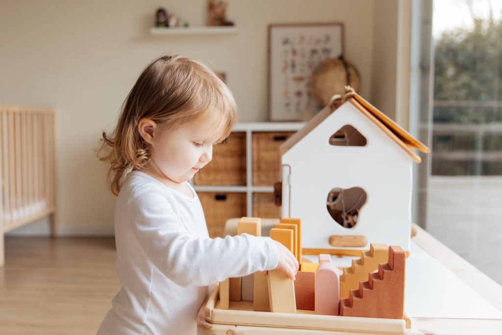 Cute little girl playing with wooden blocks at table near window at home