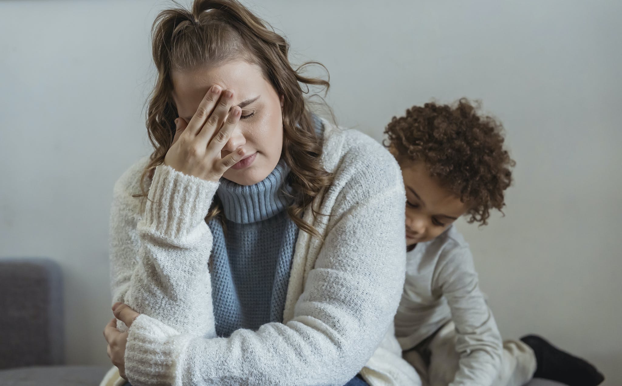 Frustrated mother with hand on forehead and closed eyes sitting near African American son near wall in room at home