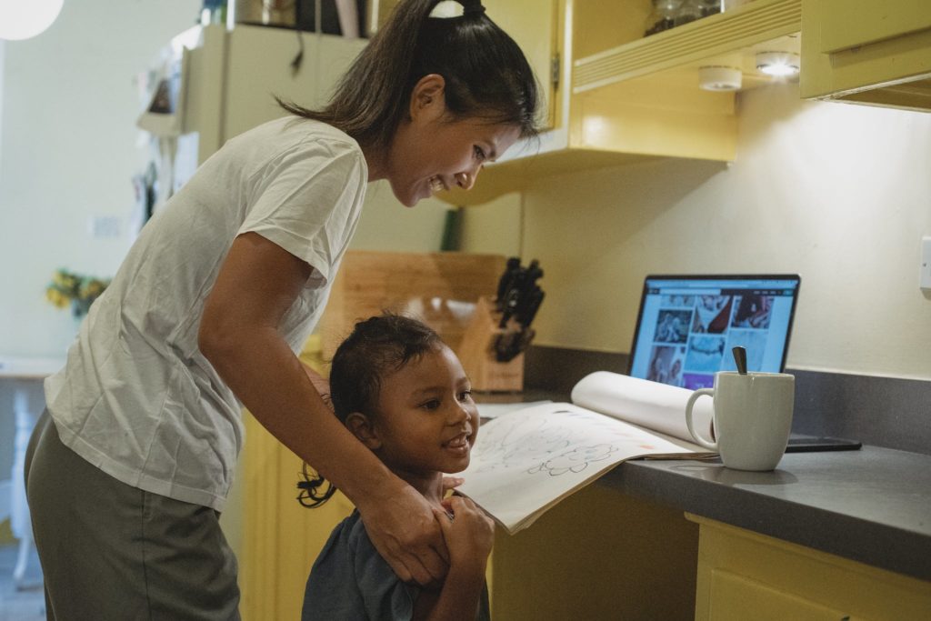 Side view of happy young Asian female freelance in casual clothes teaching little daughter to draw while working remotely on laptop at home