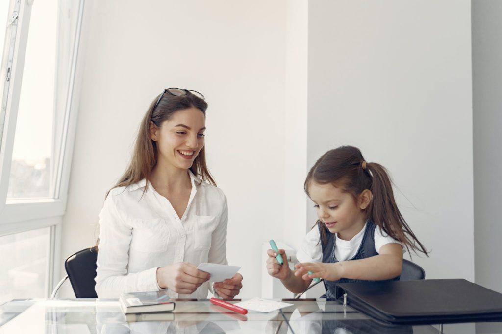 Smiling young female tutor explaining lesson for little girl and helping write while learning at table in modern light workplace