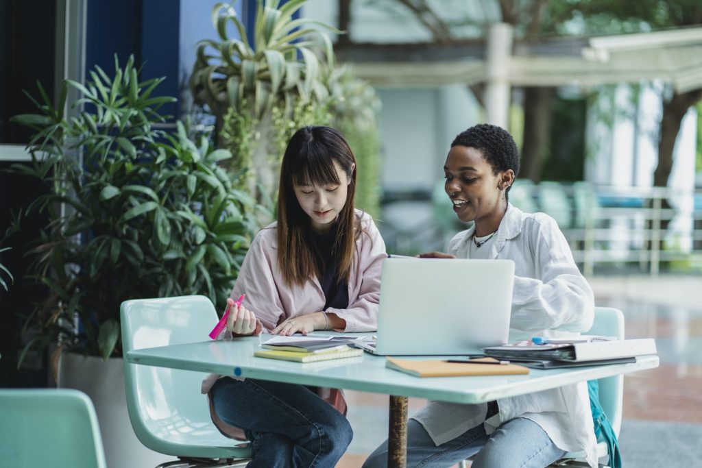 Young content diverse girlfriends talking while studying together at table with netbook in daylight