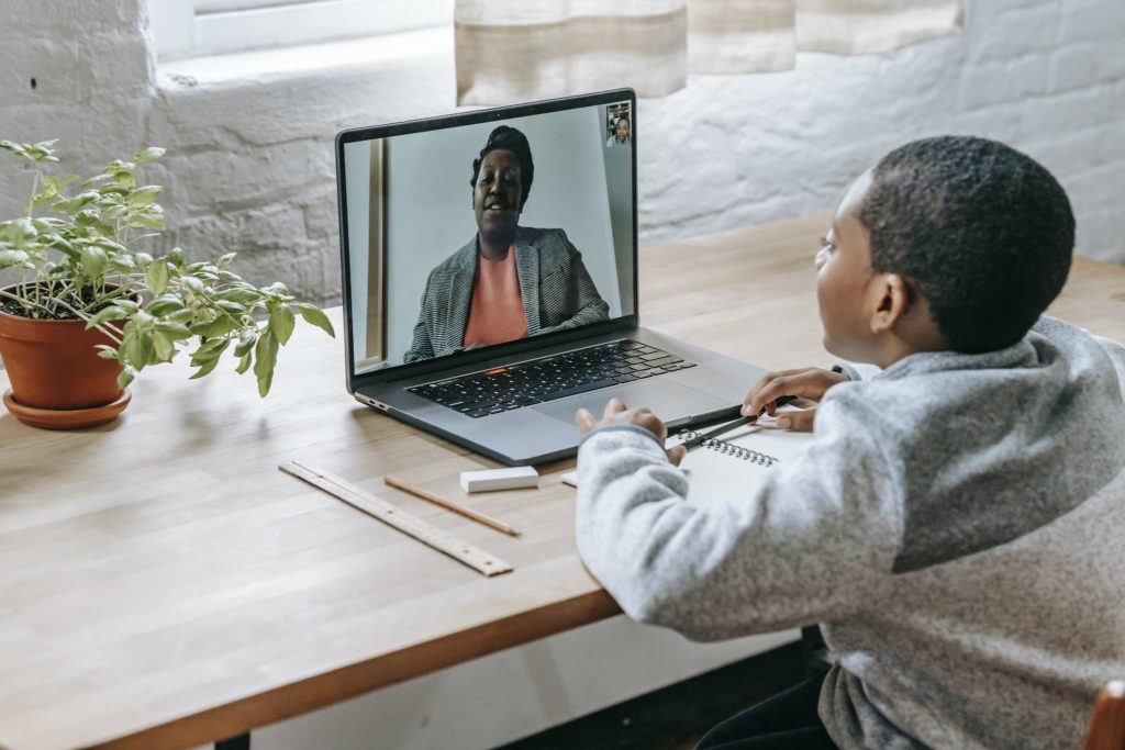 African American schoolboy sitting at desk with school supplies and laptop while looking at screen during online lesson