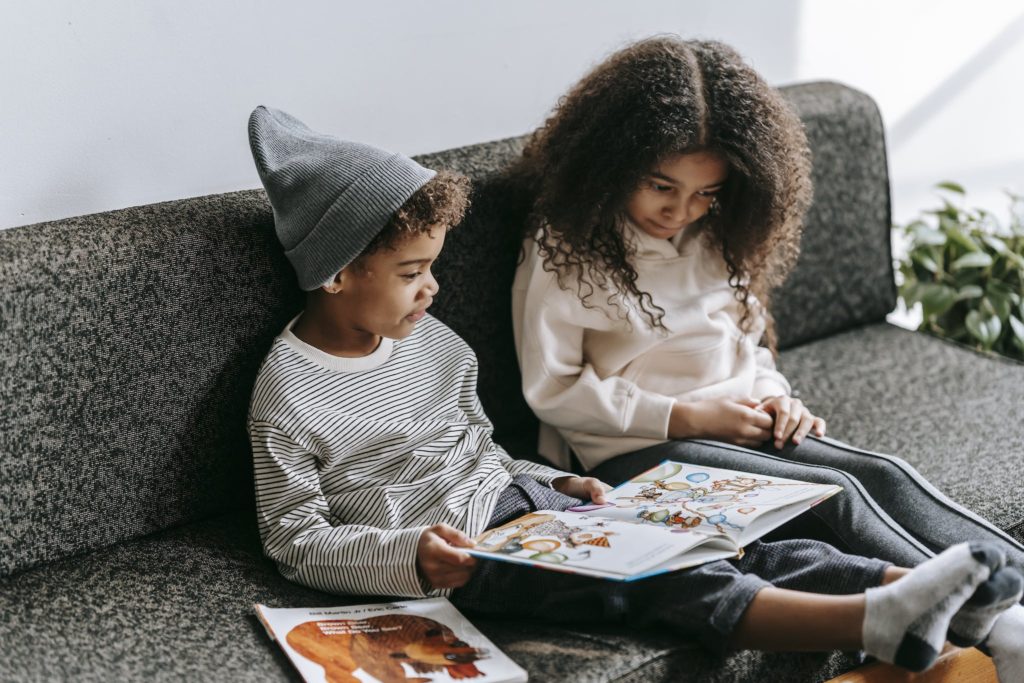 Clever African American brother and sister in trendy outfits reading interesting story with pictures in book on sofa