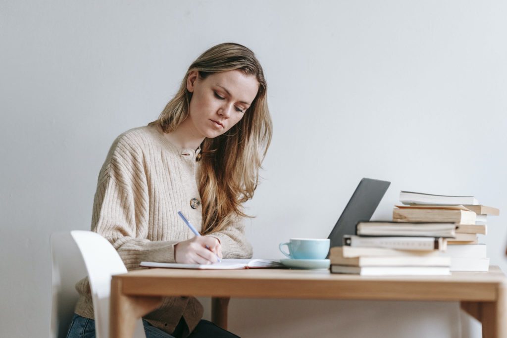 Concentrated female taking notes in planner near heap of many books and cup of strong coffee on white background