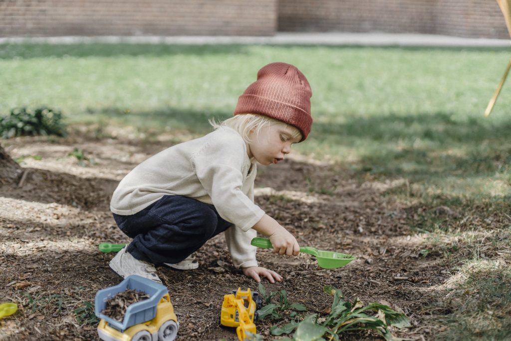 Side view of little boy in casual clothes and brown hat playing with plastic toys in backyard