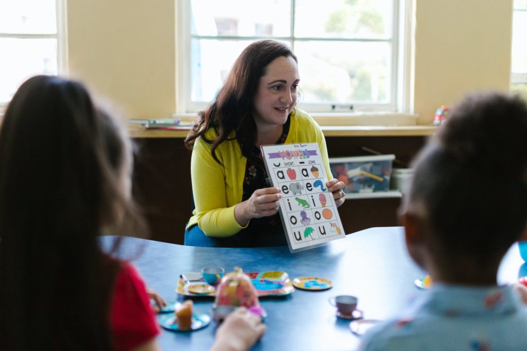 Woman in Yellow Long Sleeve Shirt Showing Letters on a Book to Kids